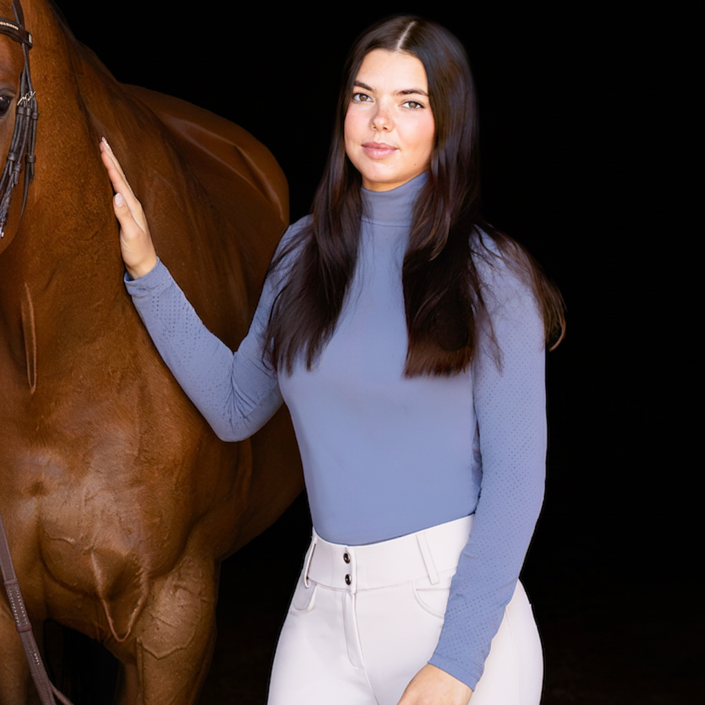 Three women standing with a brown horse in an outdoor arena at night, wearing equestrian apparel including black boots, breeches, and a white jumpsuit.